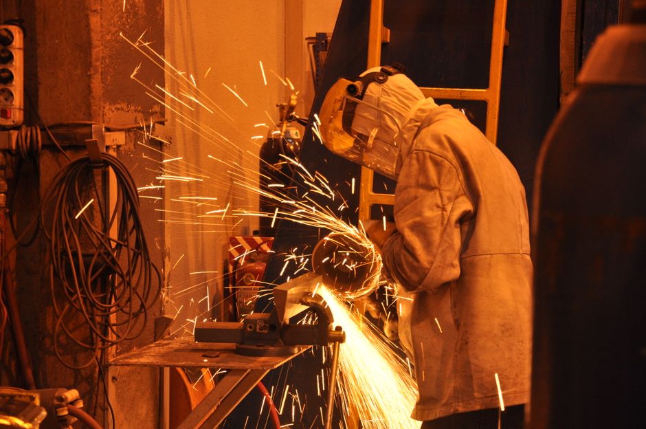 A worker in protective gear uses a grinder in an industrial setting. Sparks fly in the indoor workshop. (Photo: Uğur Bozdoğan / Pexels)