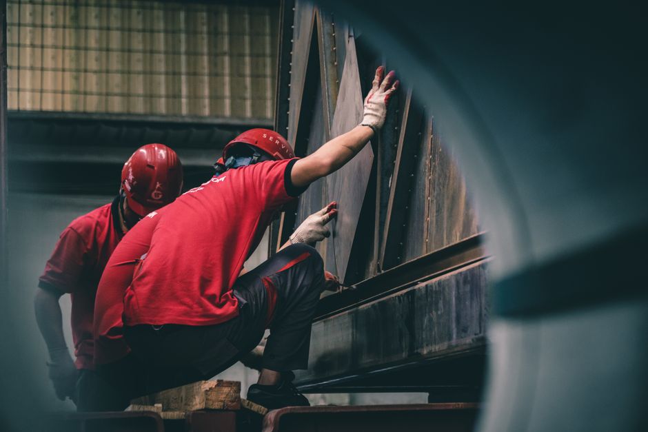 Two workers in red helmets assembling metal components inside a factory. (Photo: Hoang NC / Pexels)