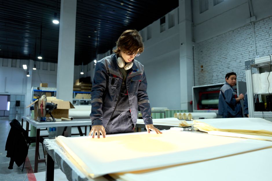 Worker in uniform examining sheets in a brightly lit factory environment, focusing on quality. (Photo: Ruslan Alekso / Pexels)