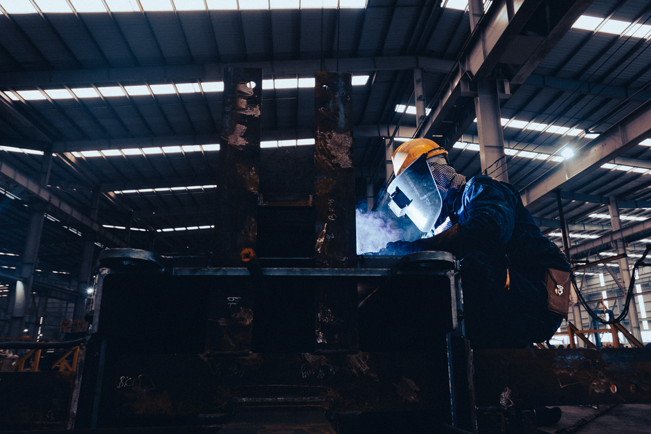 A welder wearing protective gear works on metal in a dimly lit industrial workshop. (Photo: Hoang NC / Pexels)